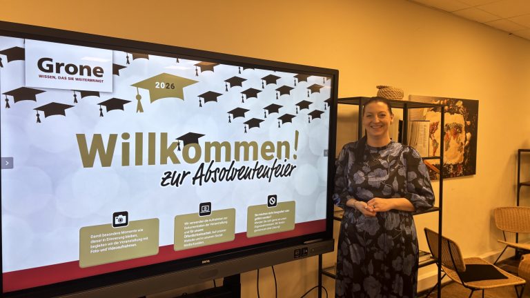 Das Bild zeigt die Schulleitung der Grone Wirtschaftsakademie Anna-Carina Gehm neben einem Bildschirm mit der Aufschrift "Willkommen".